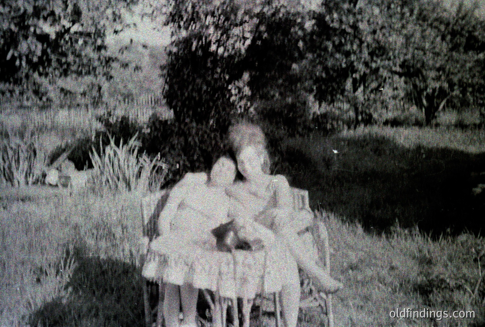 Three young girls in dresses gather near a small, wrought-iron table and chairs within a grassy, wooded area. The image has visible grain and characteristics of vintage film photography. Likely a candid snapshot, showcasing childhood and leisure. Approximate era: 1950s-1970s.