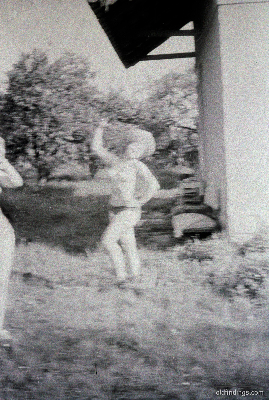 A candid snapshot captures a child mid-motion, seemingly dancing or playfully jumping. The scene is framed by a building overhang, suggesting an outdoor porch or veranda. The grainy texture and style indicate a likely 1960s or 1970s origin. A blurred background hints at a yard or garden setting. A charming, informal moment.