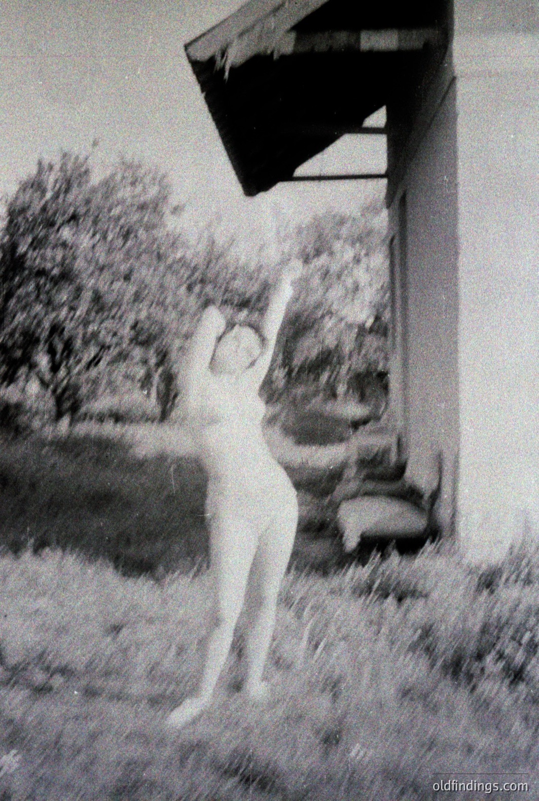 A figure stands under a projecting roofline, arms raised. The scene appears to be an outdoor courtyard or patio with tall grass and foliage visible. Likely a 1960s-70s snapshot, possibly from a resort or residential setting. The high-contrast image has a vintage aesthetic.