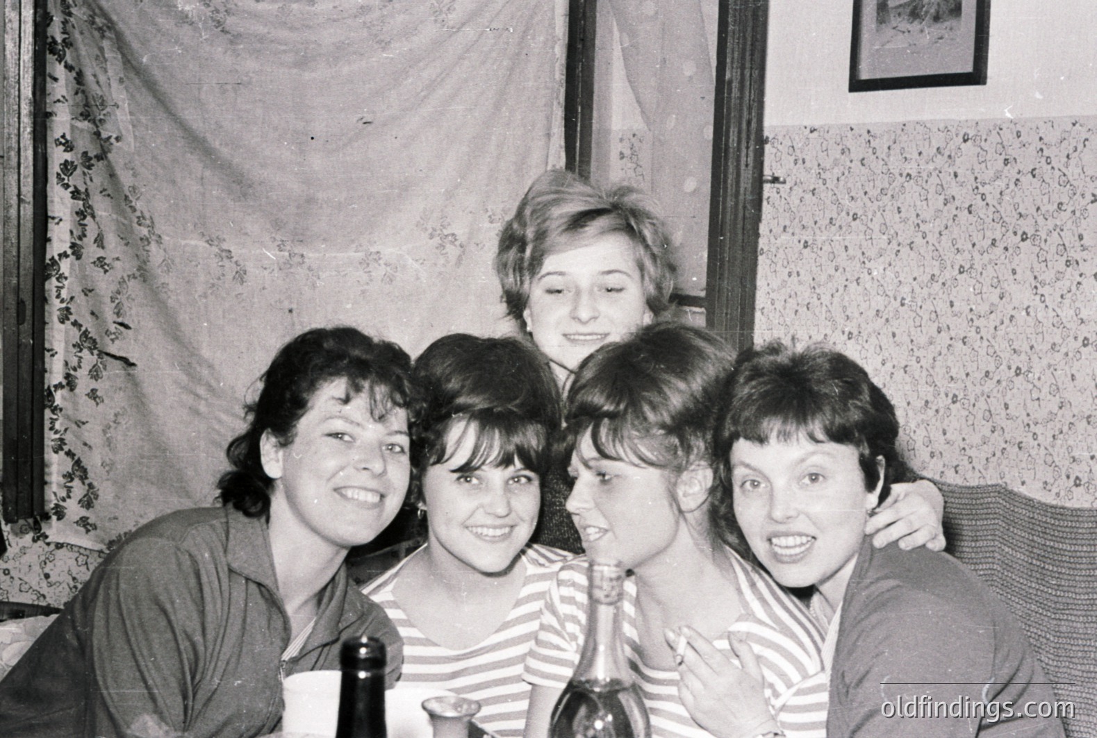 A group of four women, smiling and closely positioned, appear in a domestic setting. They are wearing striped tops and casual clothing. Likely mid-1960s, the photo showcases a candid, intimate moment. Textured wallpaper & draped fabric suggest a modest interior. Valuable for documenting social life & fashion.
