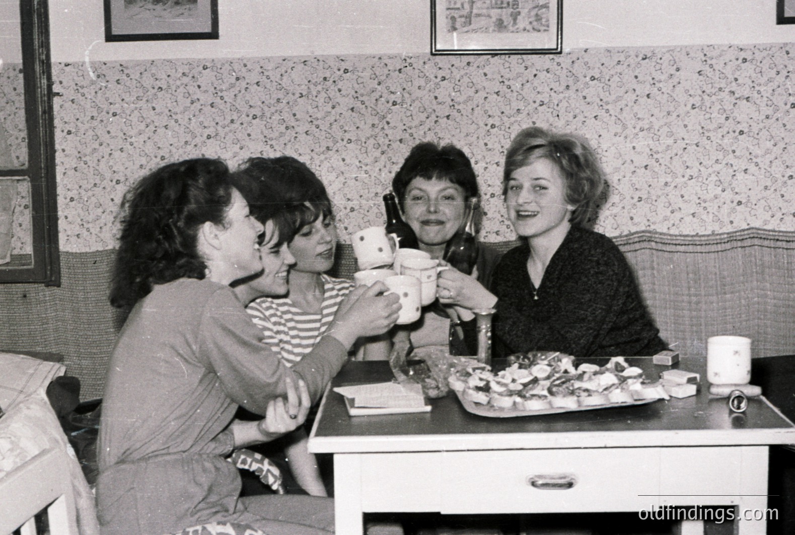 Four women toasting with mugs in a vintage cafe setting, likely 1960s-70s. Decorative wallpaper, a small table with pastries, and a booth create a cozy, intimate atmosphere. Candid moment of joy and camaraderie. Ideal for design or nostalgia projects.