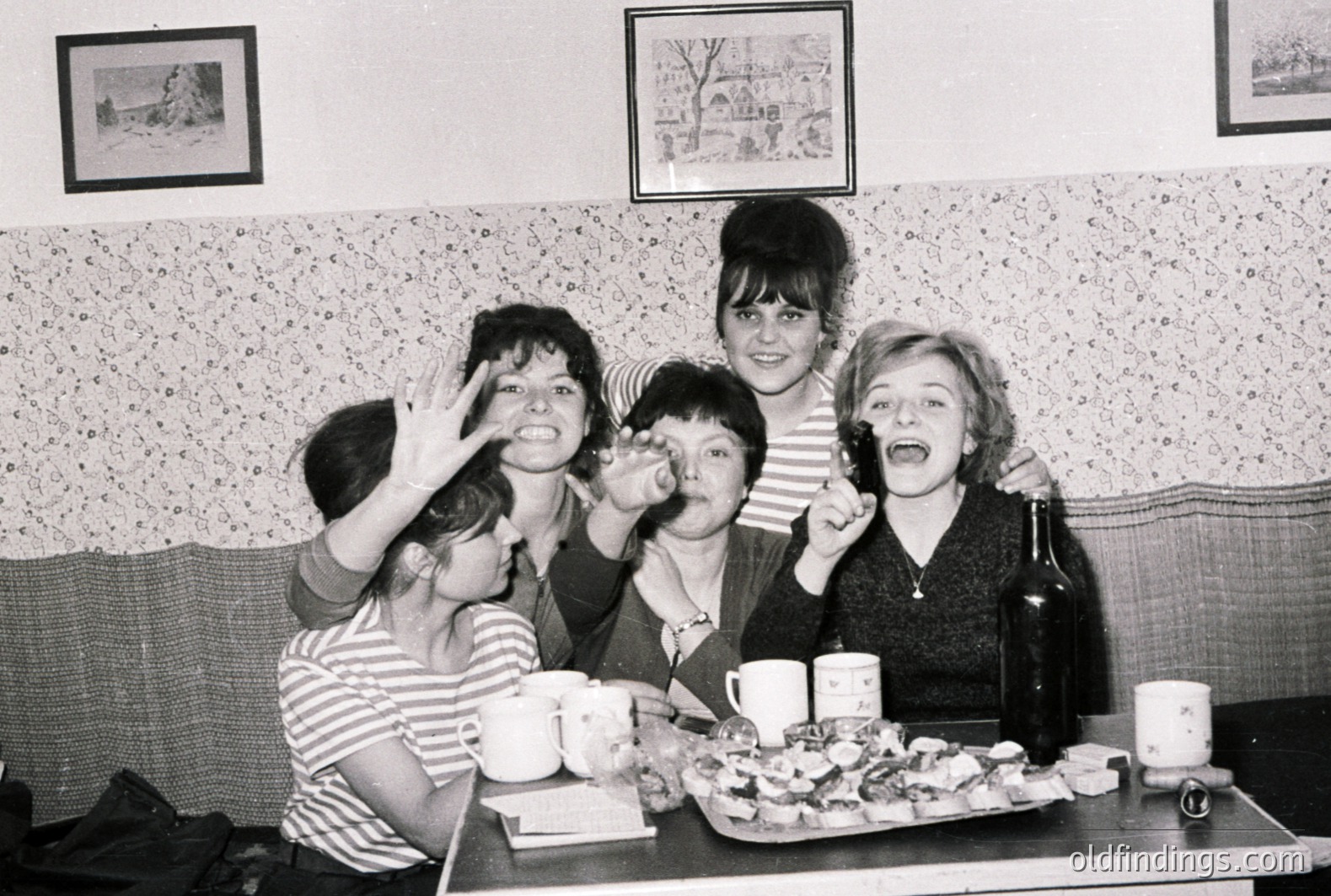 Five young women, styled in 1960s fashion, playfully pose within what appears to be a cafe or diner setting. Decorated wallpaper and framed pictures are visible. A tiered serving tray holds small cakes or pastries, accompanied by coffee cups and a bottle of wine. A candid moment, capturing youthful exuberance.