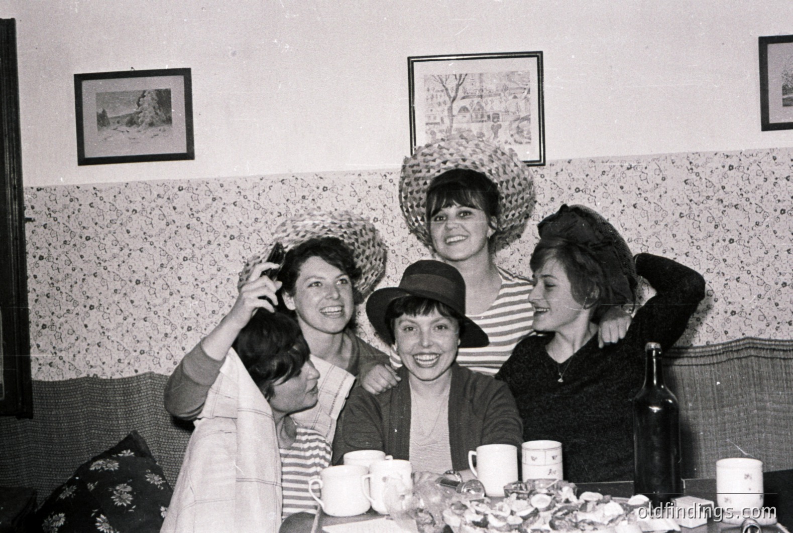 Five young women playfully pose, adorned with various straw hats. A table laden with cups, a wine bottle, and a plate of marshmallows adds a casual, convivial atmosphere. Likely a snapshot from the 1960s or 70s. Interior decor reflects a mid-century aesthetic. A charming, candid moment.