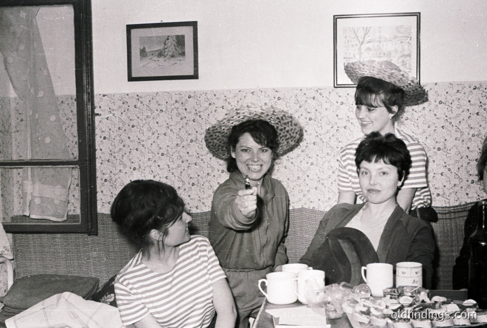 Four women socialize in a richly patterned interior; two wear striped tops & straw hats. A table with teacups & pastries sits between them. The scene suggests a casual gathering, likely a home or small cafe. Appears to be .