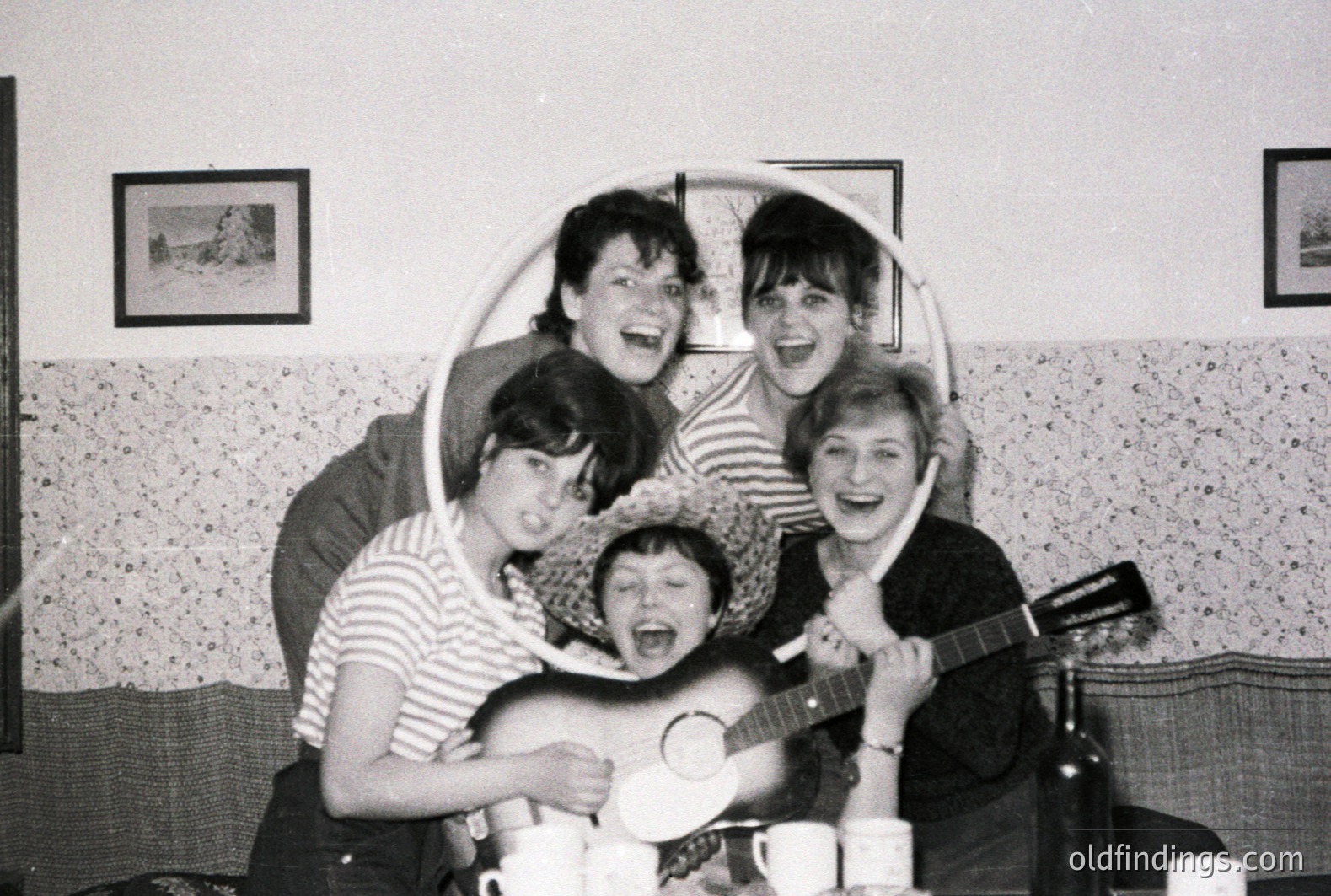 A group of five young adults playfully pose for a casual portrait. Seated amongst teacups, one plays a guitar, another dons a straw hat. Visible architectural details suggest a 1960s interior. Likely a candid moment of camaraderie.