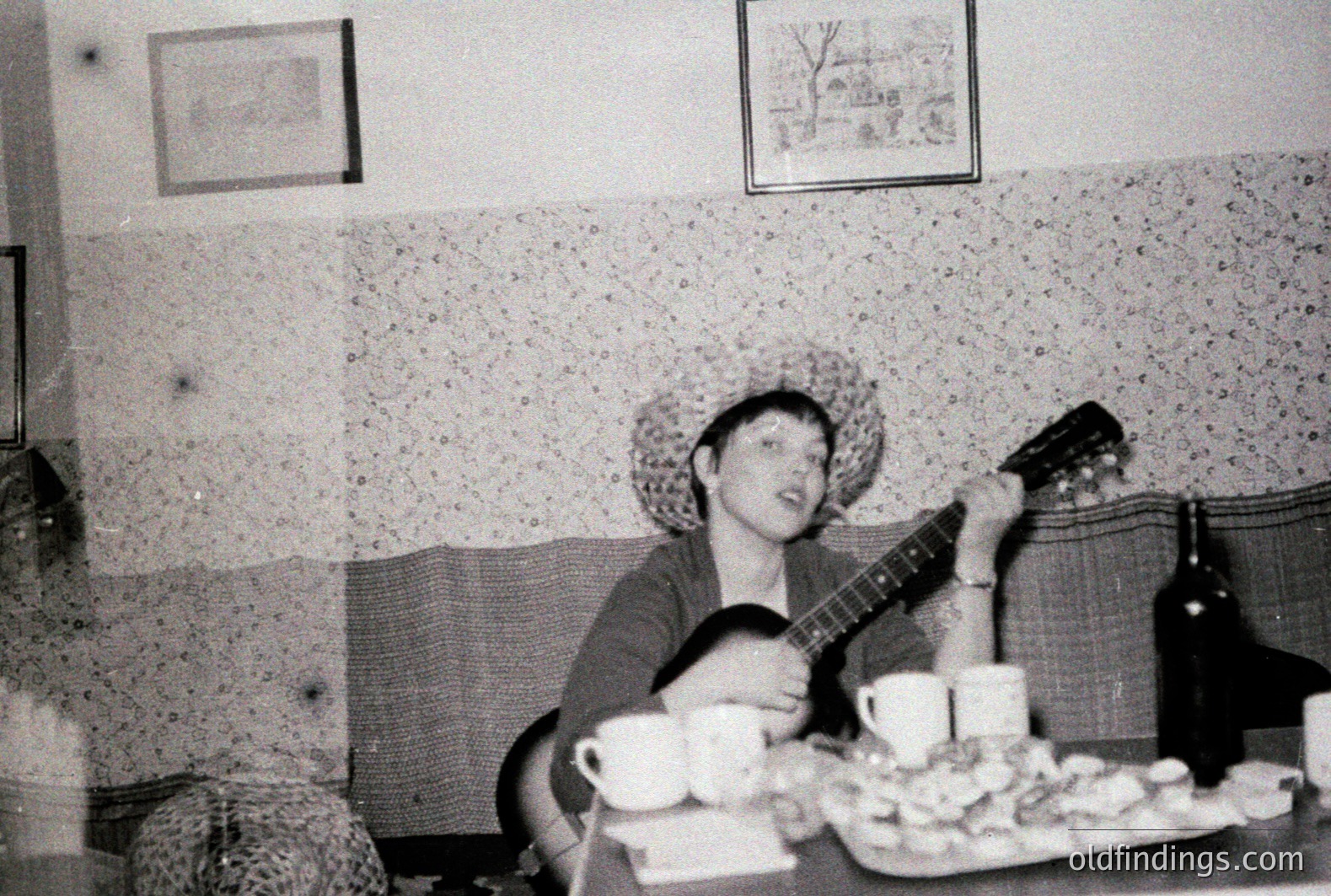 A young person seated at a table, playing an acoustic guitar. Visible are several cups and a platter of what appears to be fruit or snacks. The backdrop features textured wallpaper and a framed picture. Likely a casual, domestic scene. Possible 1970s aesthetic.