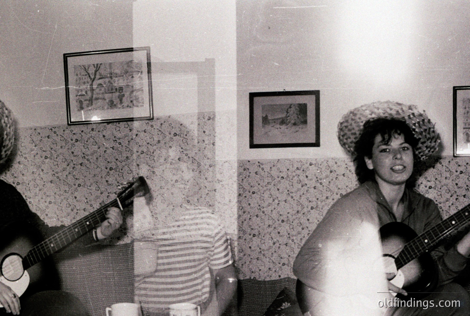 A group gathers for an informal musical gathering in a room featuring textured walls and framed landscapes. A person with a straw hat plays a bass guitar while another plays an ukulele. Casual attire suggests a relaxed, intimate setting. Likely a 1970s social or leisure moment.