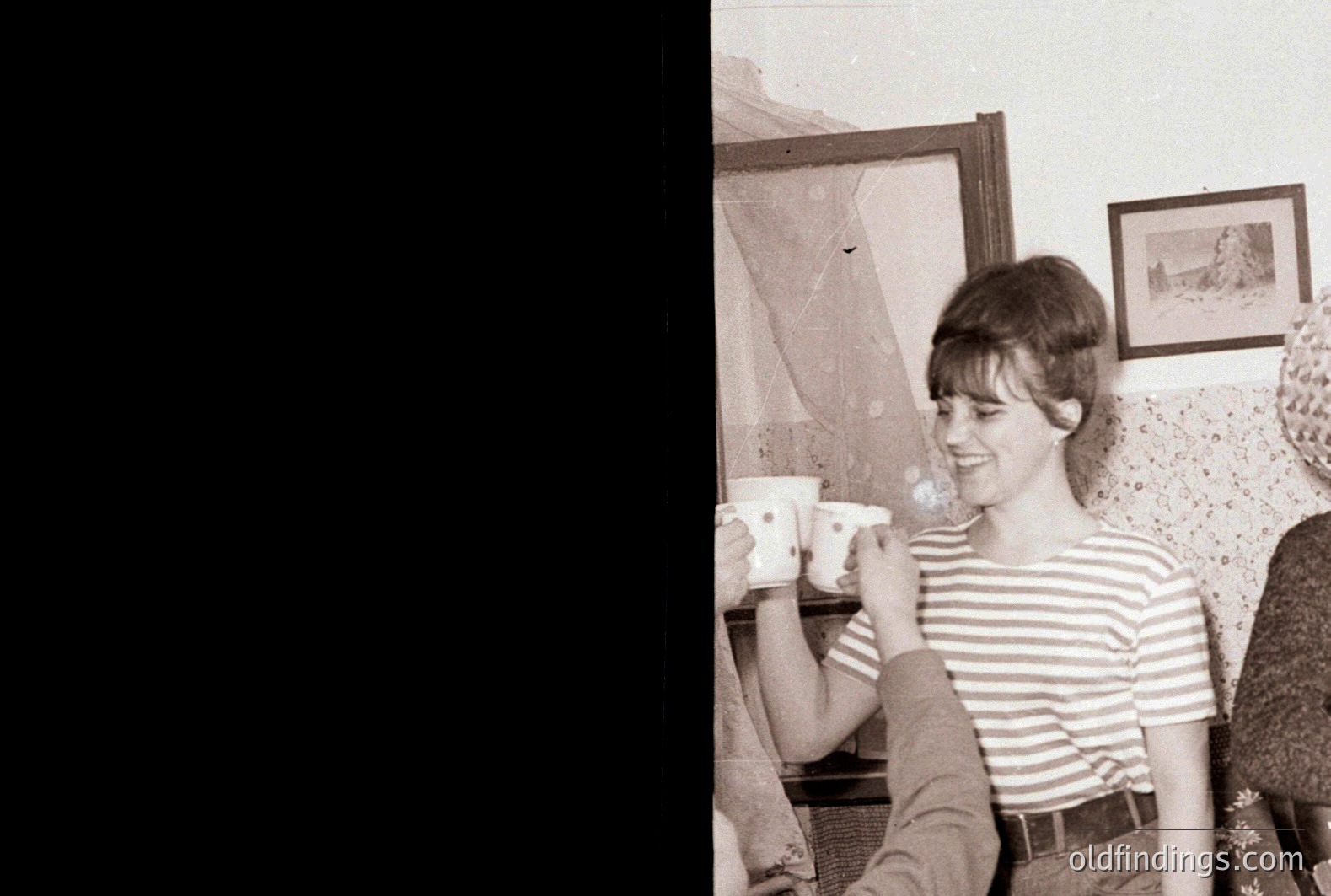 A young woman with a short bob haircut smiles while participating in a tea ceremony. She wears a striped top and appears relaxed. Interior setting: framed artwork on the wall and floral patterned wallpaper. Likely a domestic or informal gathering.