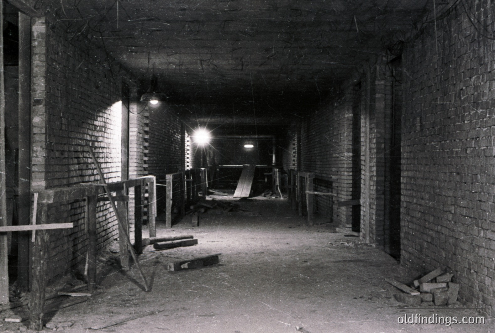 A long, dimly lit brick corridor or tunnel. Construction scaffolding and lumber are present, suggesting ongoing or recent work. The walls are brick, showing signs of age and wear. Single light illuminates the scene, casting long shadows. Likely a building interior, industrial or utility space.