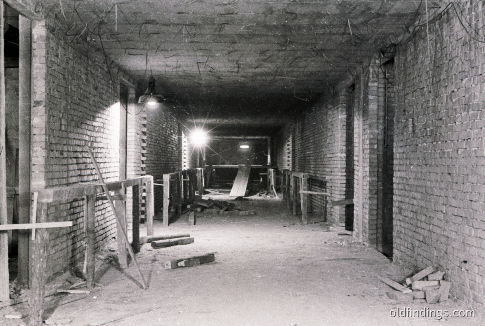 A stark, long corridor with exposed brick walls and a low ceiling. Construction scaffolding and timber supports suggest ongoing work. Sparse lighting reveals a dusty, unfinished space. Possible industrial or utility area, circa 1950s-1970s. Useful for architectural reference or stock photography.