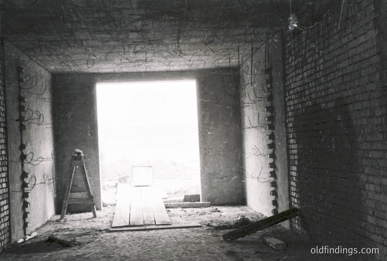 An unfinished brick interior reveals an exterior view through a doorway, suggesting construction or renovation. A wooden plank bridge spans across a void, with scaffolding visible. The scene appears dated, likely 1960s-1970s, judging by the photographic style and construction techniques. Industrial, raw aesthetic.