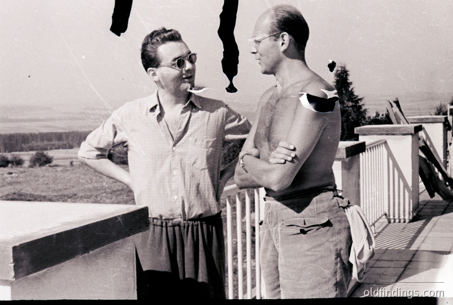 Two men converse on a balcony overlooking a forested landscape. One smokes, both wear glasses. Casual summer attire suggests a relaxed setting. Likely mid-20th century, possibly 1950s or 60s, based on style. Architectural details indicate a hillside dwelling. Potential for design reference or nostalgic appeal.