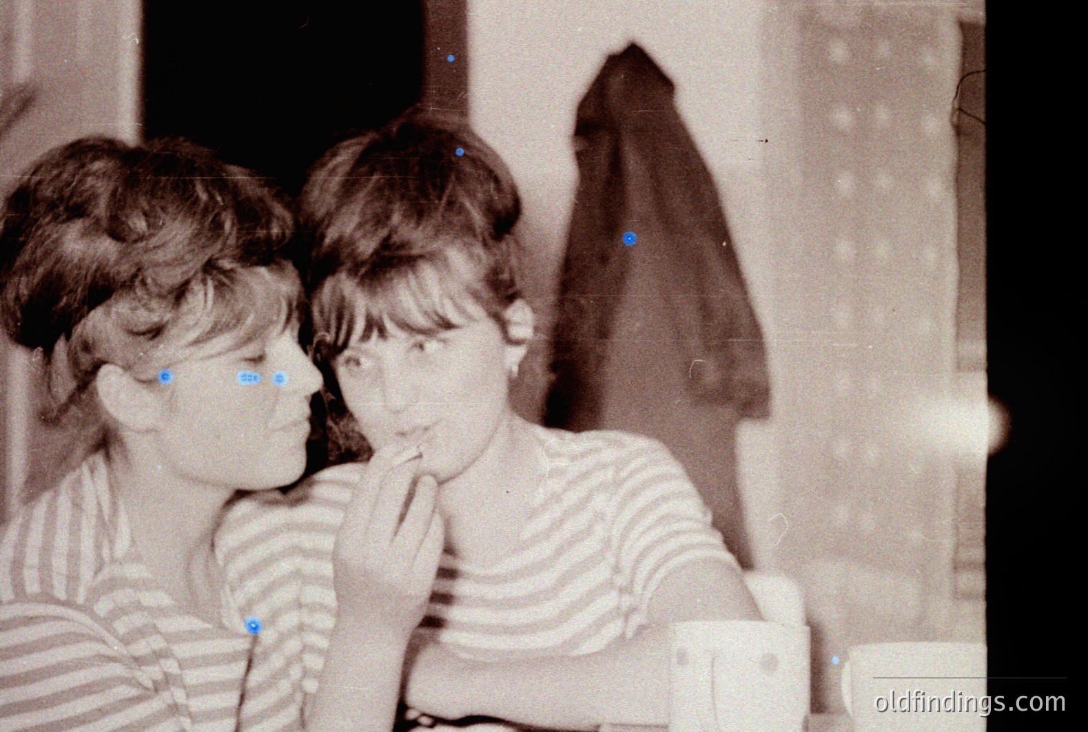 Two young women, striped tops, close proximity. One gently touches the other's face. Interior scene, likely a home, with a dark curtain visible. Possible 1960s or 70s amateur snapshot. Soft focus characteristic of older film photography. Potential stock photo reference for vintage style.