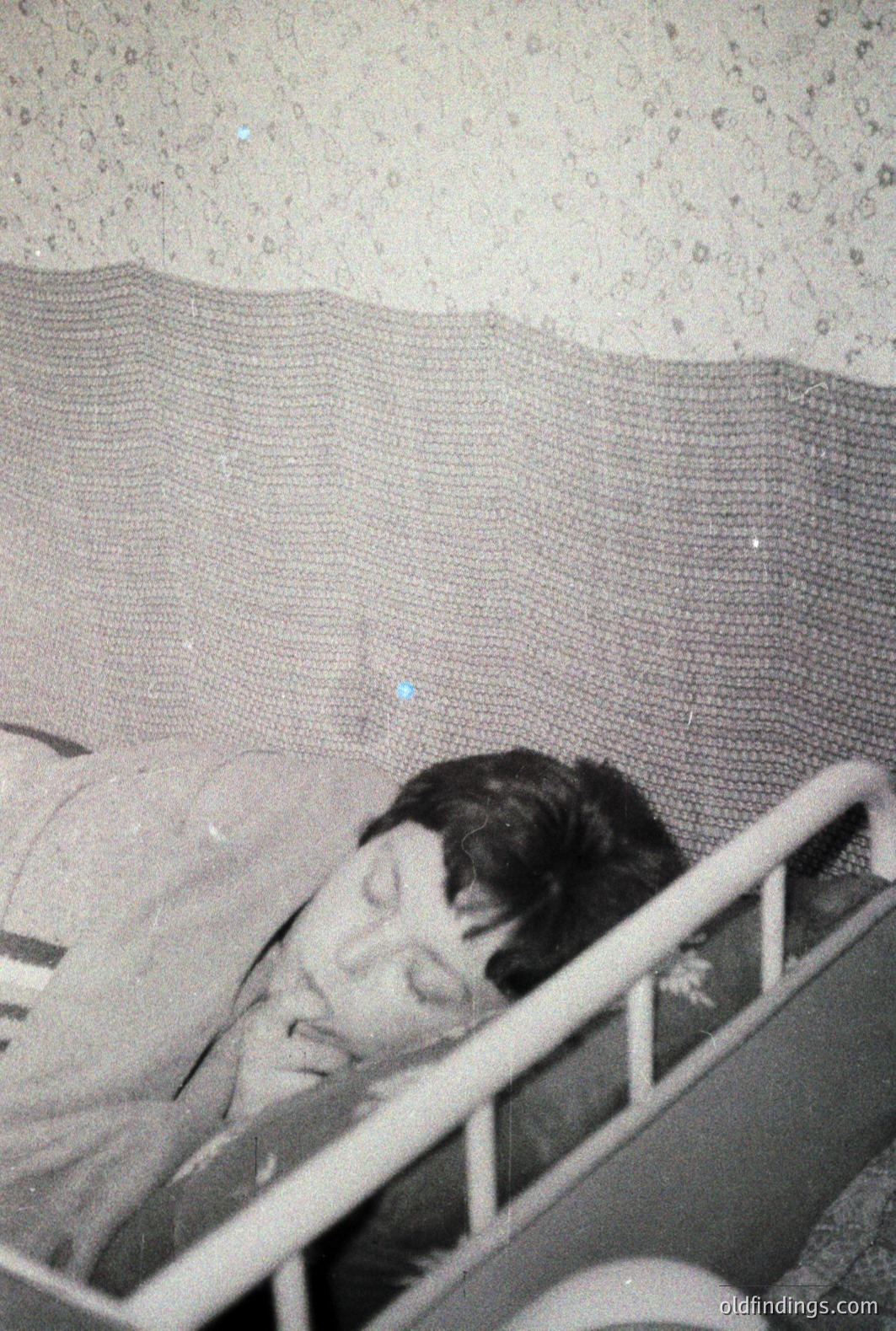 A young child sleeps in an iron crib with a textured, woven blanket. The grainy image suggests a vintage photograph, potentially from the mid-20th century. The crib's design points to a period of simple, functional furniture. Likely a family or personal snapshot.