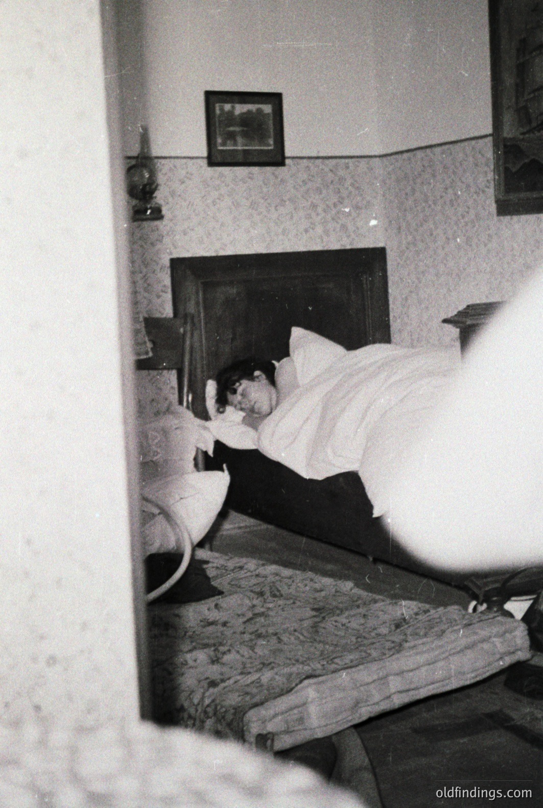 A person lies asleep in a bed with a tall, dark wooden headboard. The room’s wallpaper is patterned, and a framed picture hangs on the wall. A bedside table holds a lamp. Likely a candid, domestic snapshot, c. 1960s-1970s. Good for design reference.