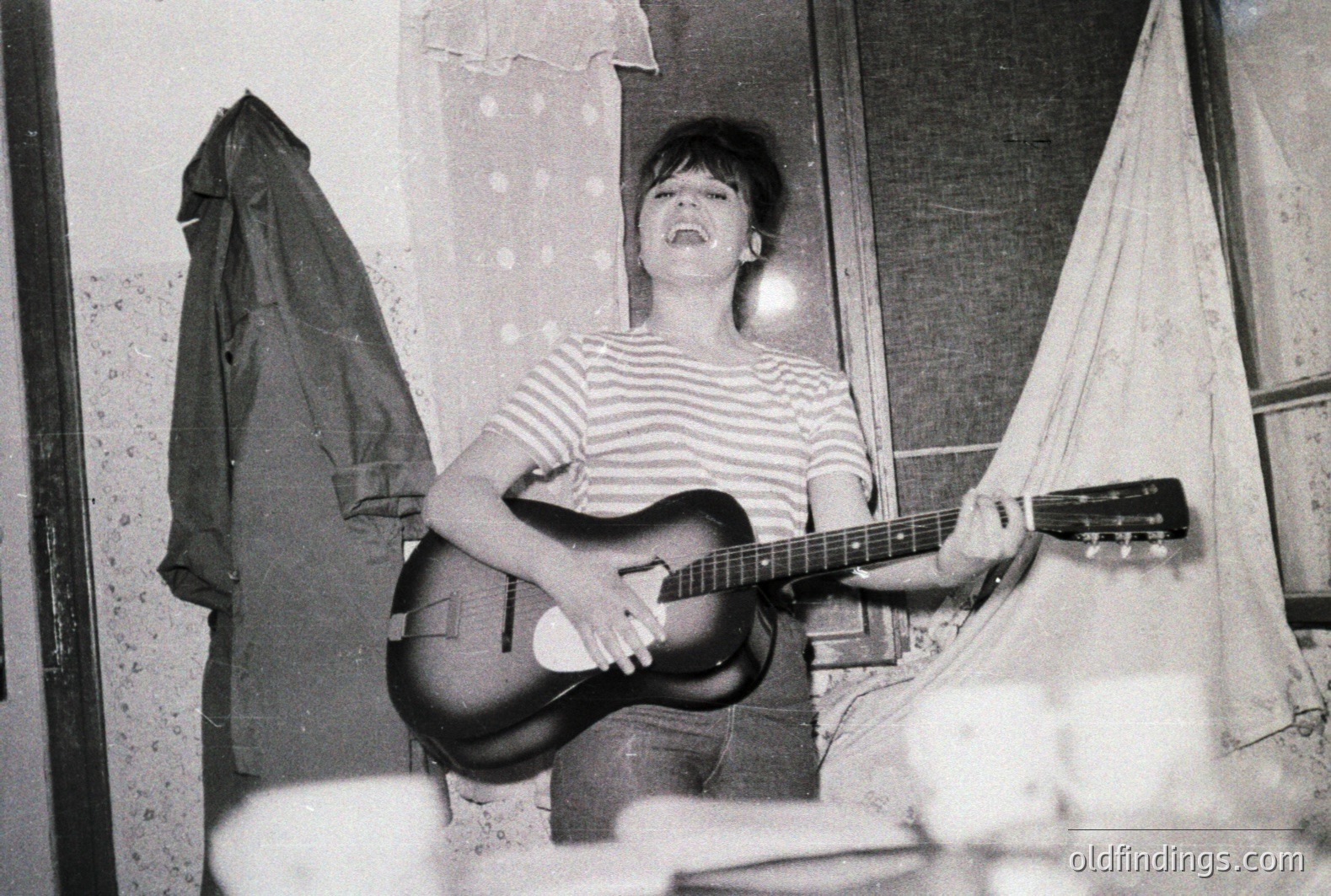 A young woman stands playing an acoustic guitar in what appears to be a sparsely furnished room. She wears a striped shirt and jeans, her expression suggesting she's singing. A coat hangs on a hook beside a window with a sheer curtain. Likely 1960s or 70s snapshot.