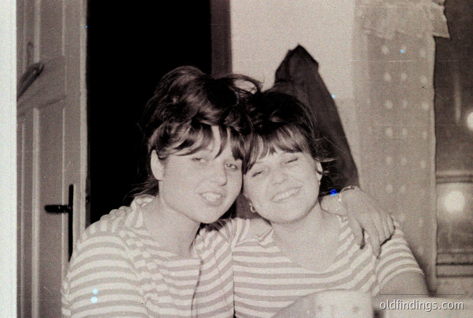 Two young women, dressed in matching striped tops, share a close, joyful embrace. The photograph, likely taken indoors near a doorway, showcases a casual, intimate moment. The style of dress and hairstyles suggest a 1970s timeframe. Strong potential for design/nostalgia reference.