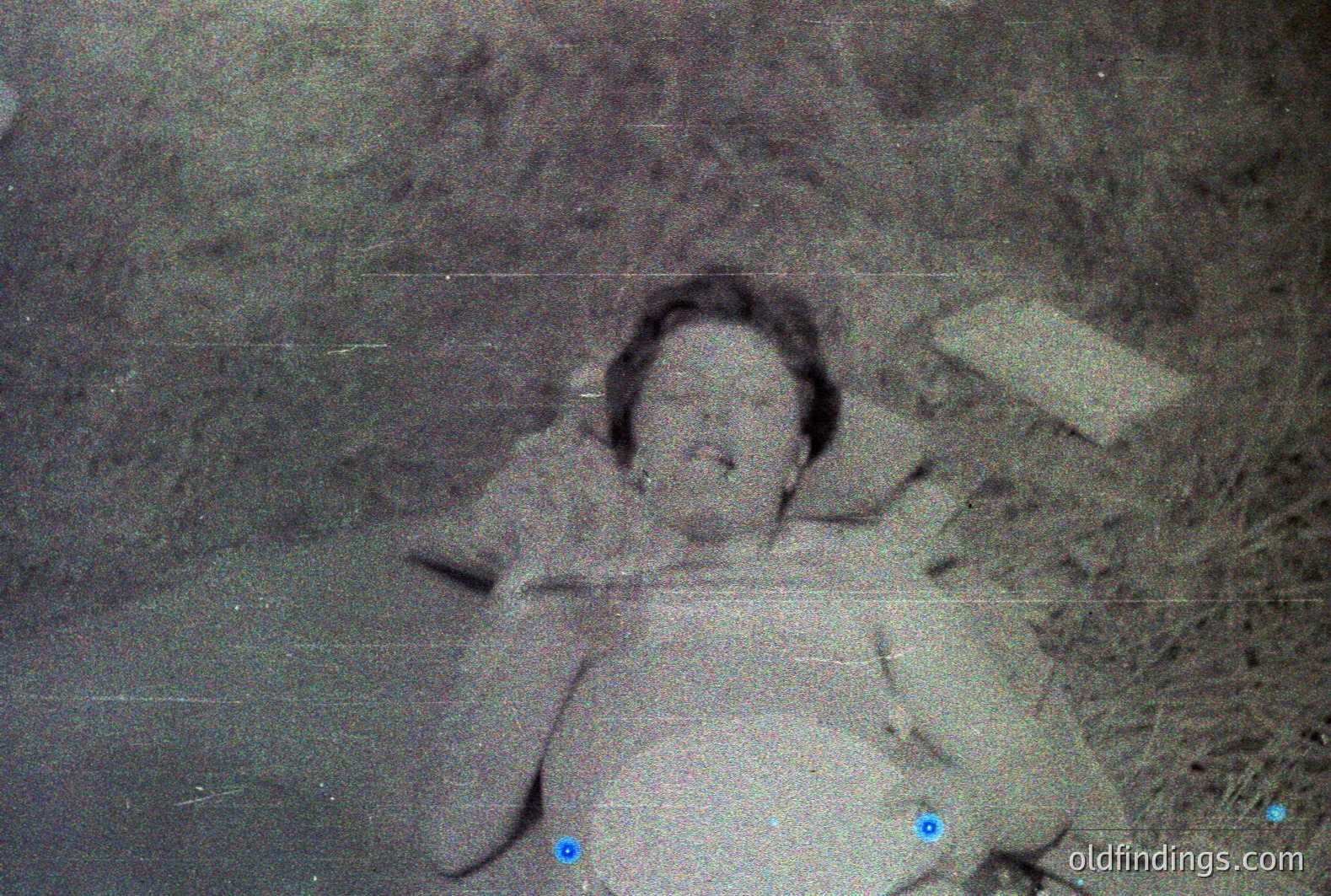 A young child lies on a bed of foliage, appearing to be outdoors. The image has a grainy, aged quality with visible film grain and scratches, indicating it's likely a vintage photograph. Shallow depth of field blurs the background. Appears to be a candid, informal snapshot.