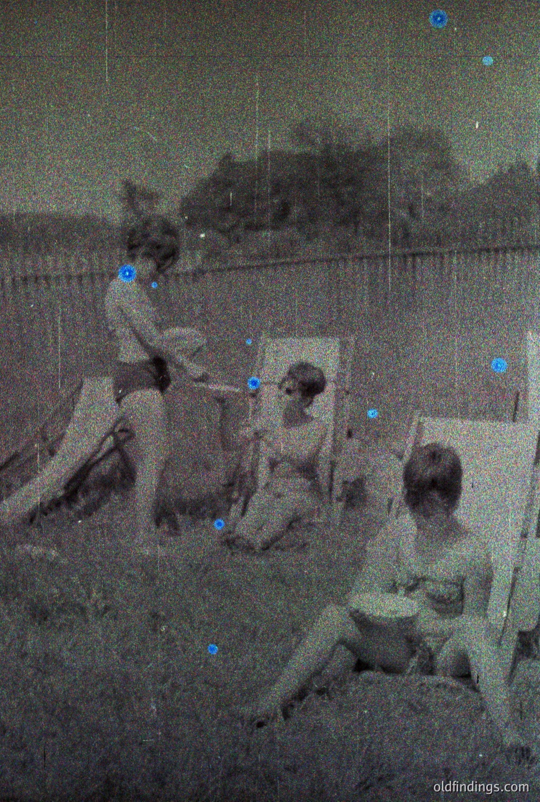 Vintage monochrome photo shows three children near a wooden fence, seemingly engaged in playful activity. One child holds a small bucket, while the others relax on the ground. Likely a backyard scene, possibly 1950s-1970s. Amateur photography evident. High archival research potential.