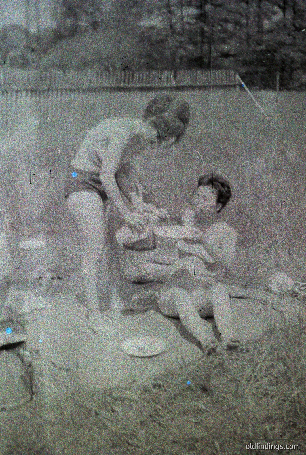 Three children play outdoors; two kneel and one stands, appearing to engage with something on the ground, possibly toys or stones. The image exhibits characteristics of a grainy, vintage photograph. Likely a candid snapshot capturing childhood leisure.