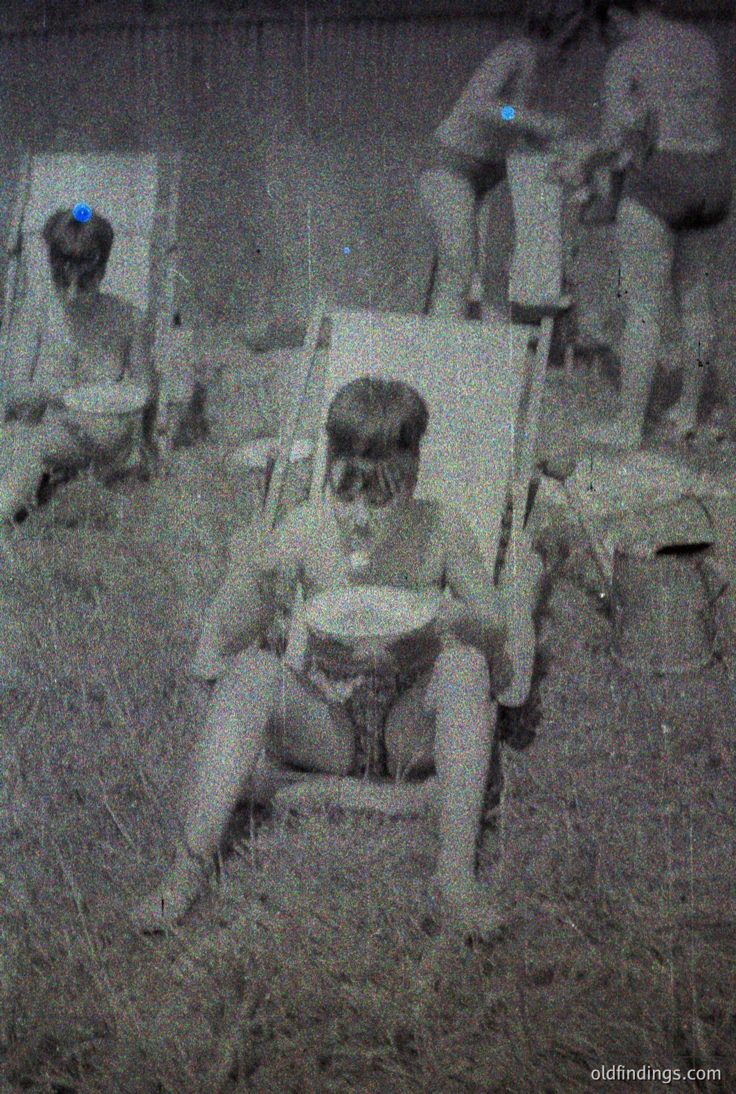 A grainy black and white photograph depicts two young boys seated in folding chairs outdoors. One boy sits facing the camera, wearing what appears to be a cap and swimwear. The other is partially obscured behind the first, also in swimwear. The setting appears to be a grassy area. Likely a family snapshot from the mid-20th century. Possibly from a vacation.