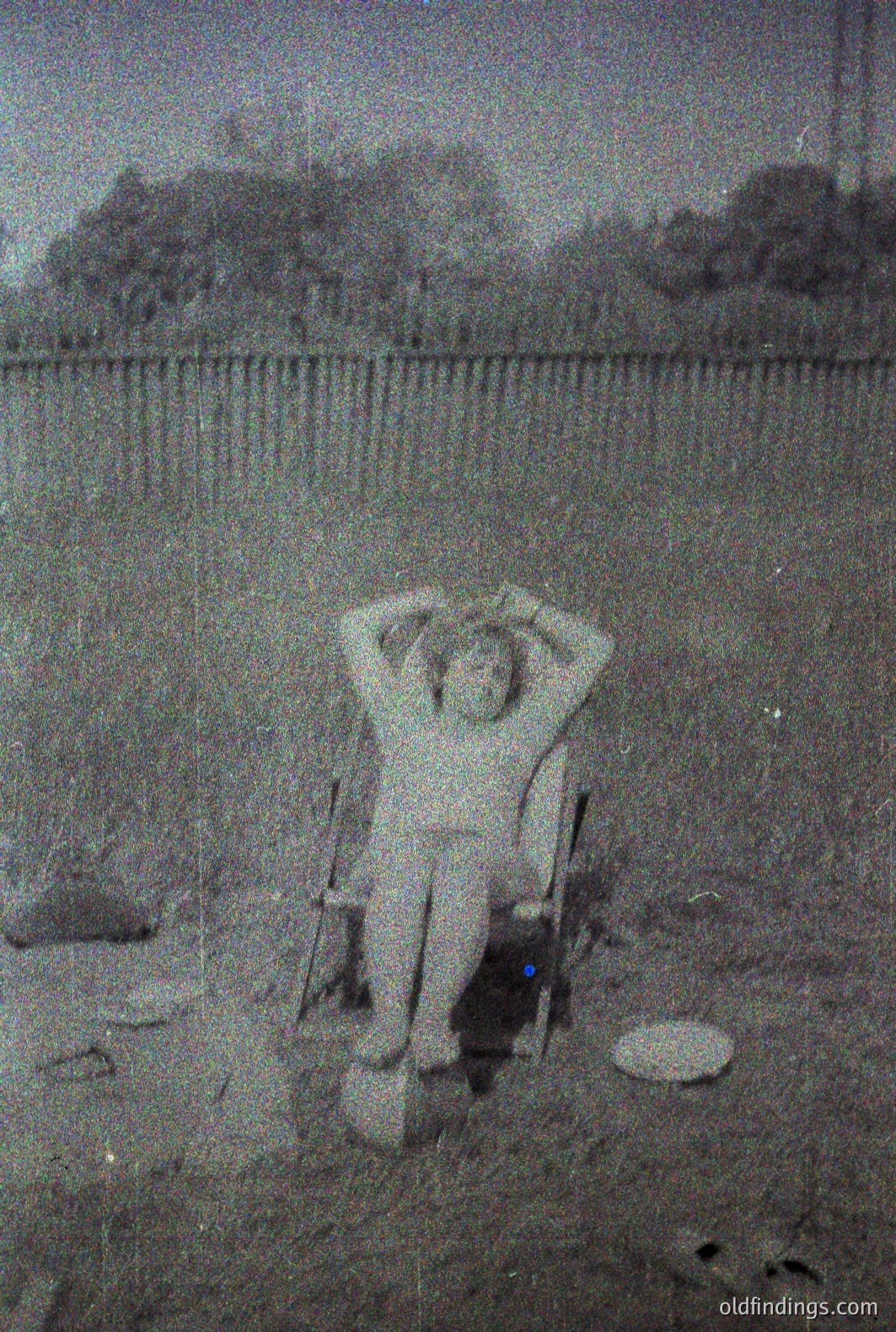 Black and white photo captures a young person seated in a folding chair, arms raised, appearing to pose outdoors. Visible foliage and an indistinct wall form the backdrop. The image exhibits visible grain and wear consistent with age. Likely a personal snapshot, possibly 1970s.