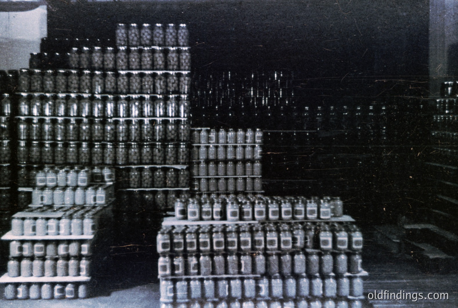 Stacked crates overflow with glass jars, likely canned goods, within a dark warehouse or factory setting. The monochrome image suggests an industrial process or storage facility, potentially from the mid-20th century. A visual record of mass production & preservation.