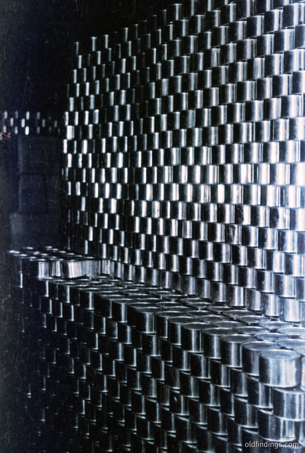 A striking view of stacked, cylindrical metal containers, likely storage drums or barrels, forms a textured wall. The arrangement creates a rhythmic pattern, diminishing in perspective. A monochromatic color palette, possibly from early color film, enhances the industrial aesthetic. Possibly 1970s. Valuable for design and industrial photography references.