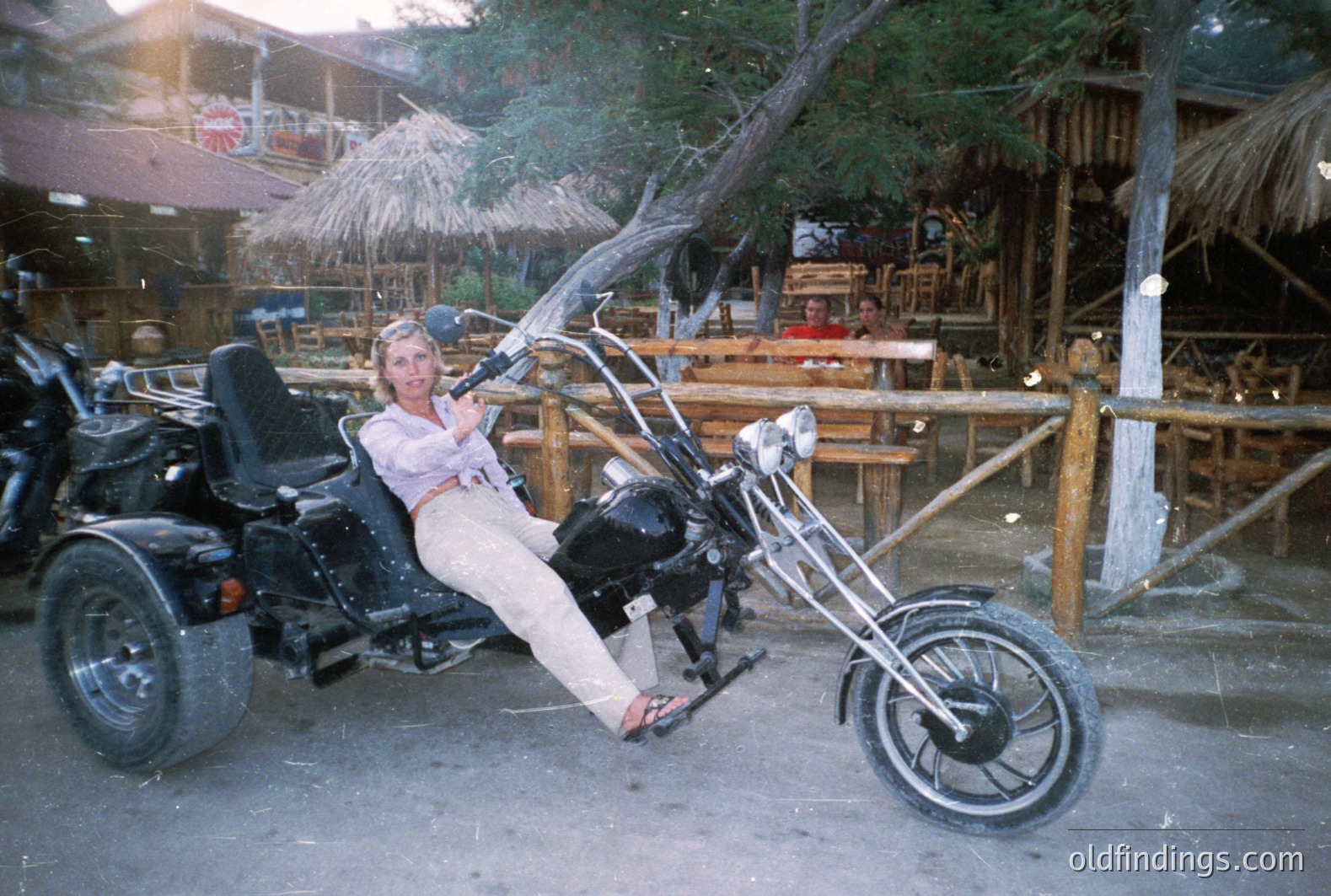 A woman sits on a custom three-wheeled motorcycle, posed casually. The background features a thatched-roof bar and patio seating, suggestive of a resort or tourist location. Appears to be a candid snapshot, possibly 1970s-80s. Likely for vintage travel or lifestyle stock.