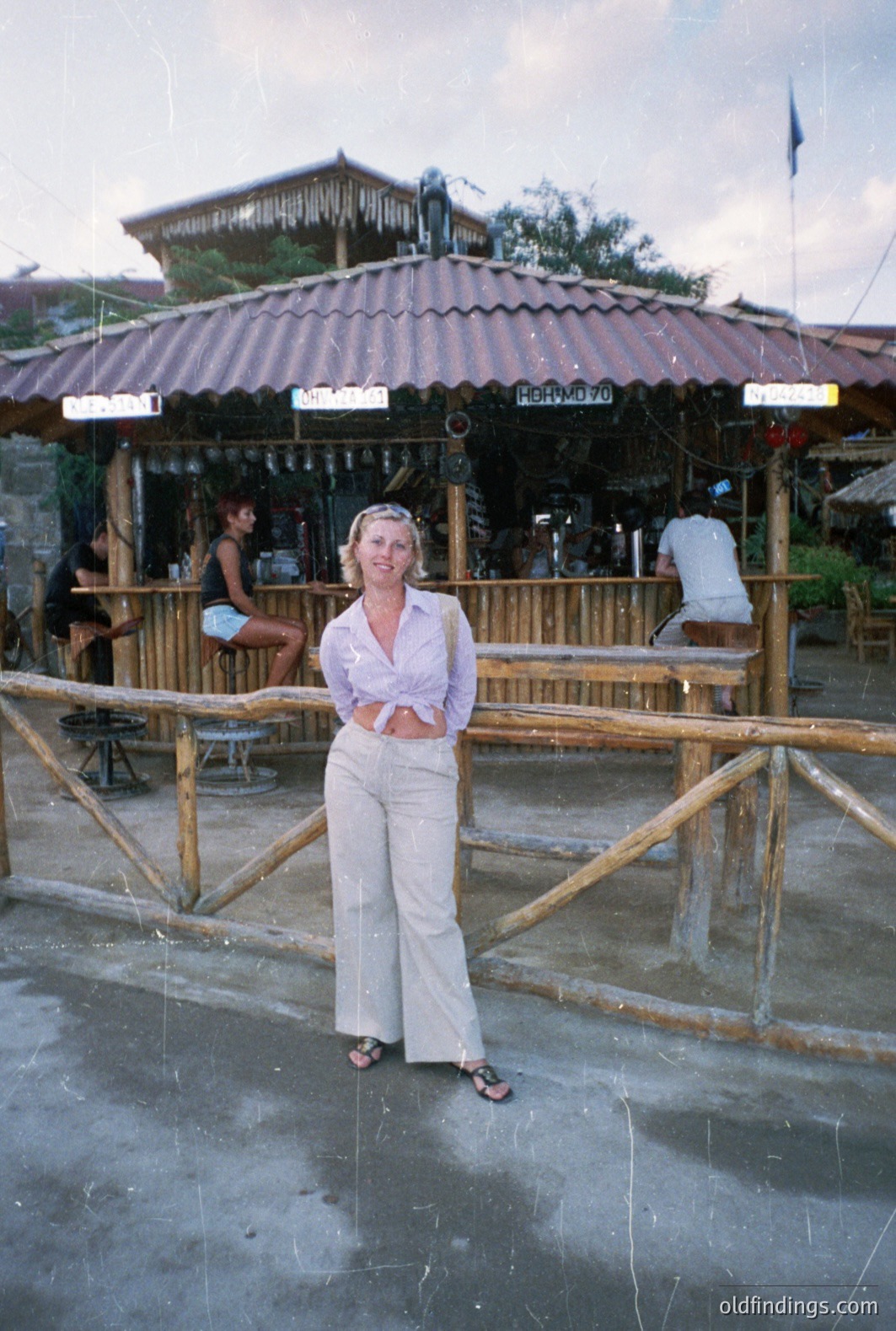 A woman stands near a beachside bar, posed for a portrait. She wears a cropped floral shirt and wide-leg trousers; sandals suggest a relaxed, vacation vibe. The bar has a thatched roof and visible signage. Appears to be a 1970s snapshot of leisure travel. Likely a coastal resort.