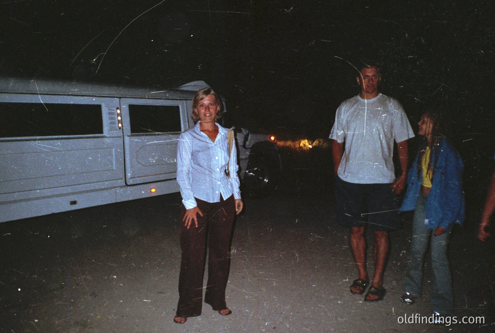 Nighttime scene: three figures stand on sand beside a large, vintage campervan, possibly at a beach. Woman in a button-down shirt & trousers, man in a t-shirt & shorts, a teen in a denim jacket. Likely 1970s or 80s casual style. Appears to be a candid vacation snapshot. Limited detail due to low light.