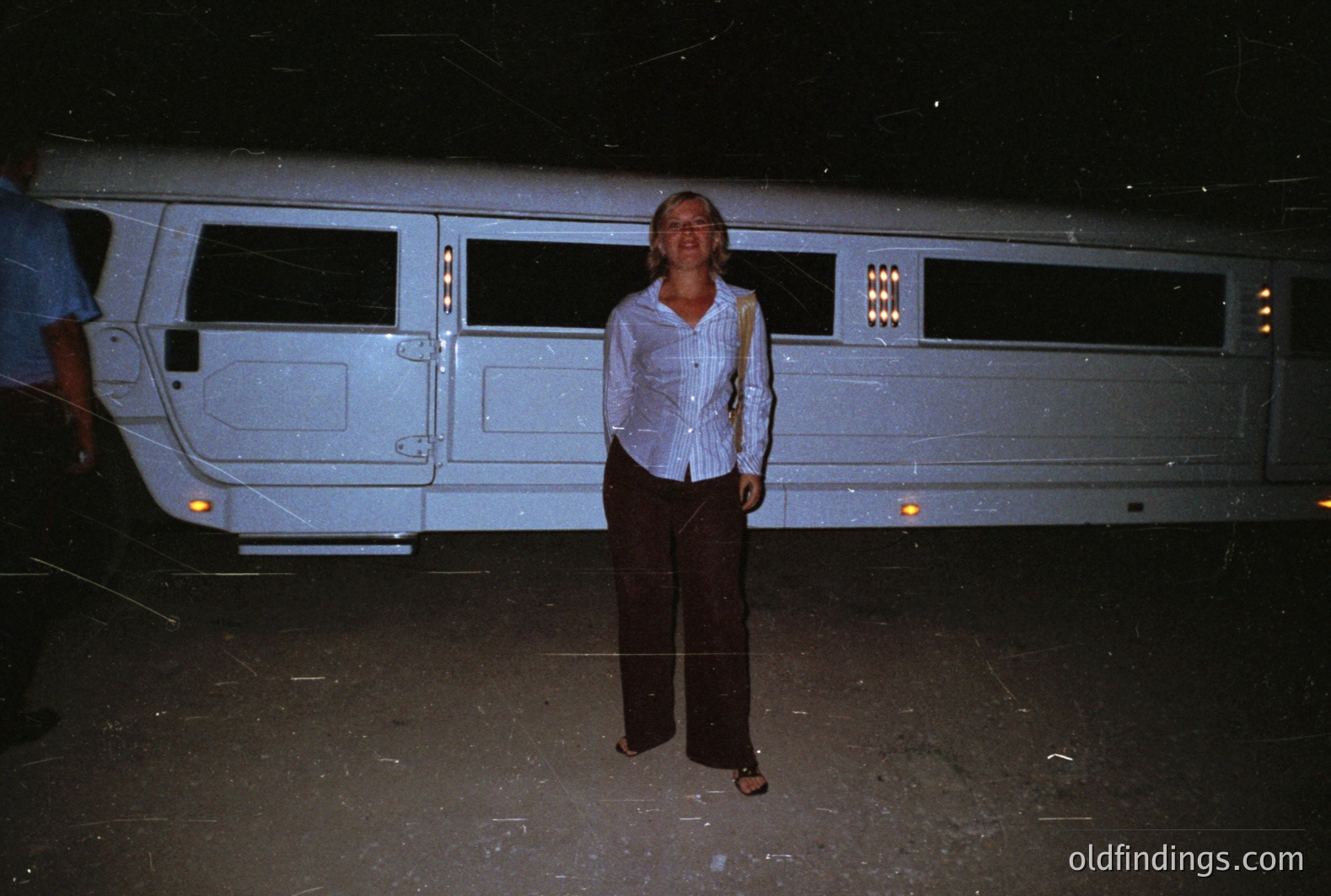 A woman stands outdoors beside a long, white stretch limousine. She wears a light-colored, collared shirt and dark trousers. The scene is dimly lit, seemingly at night, with a grainy quality suggesting film photography, possibly the 1970s or 1980s. The limousine’s extended length and interior lighting are prominent.