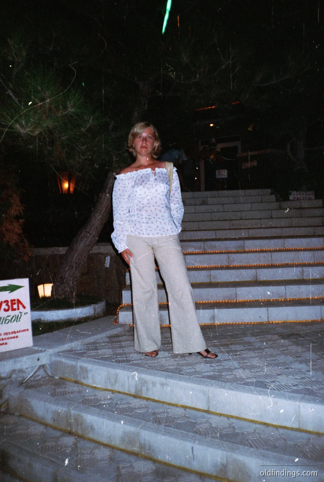 A woman stands on stone steps at night, wearing a patterned off-the-shoulder blouse and flowing wide-leg trousers. Sign visible suggests a coastal location, possibly Bulgaria. Likely taken in the 1970s or 1980s – note the clothing style & film grain. A resort or vacation setting.