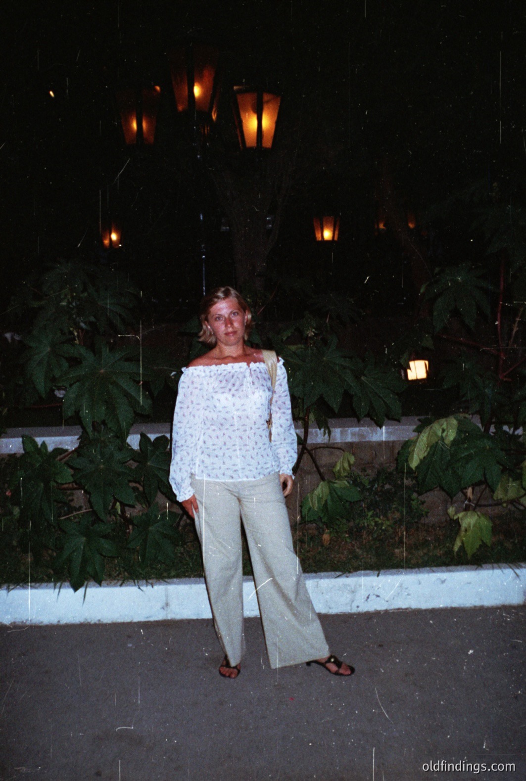A woman stands outdoors at night, wearing a patterned off-the-shoulder top and wide-leg pants. A vintage aesthetic suggests a 1970s timeframe. Background features lush foliage, and lampposts. Possibly a resort or vacation setting. A candid snapshot reflecting leisure travel style.