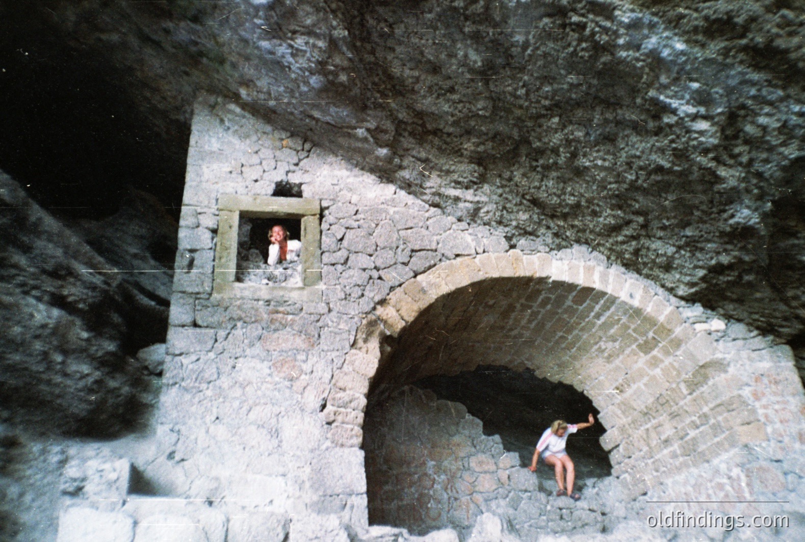 Stone archway & window integrated into a rugged, dark rock face. A figure sits within the arch, another observes from above. Likely an alpine structure, possibly a former shepherd's refuge. Appears to be a travel photograph, likely 1970s. Offers design reference for rustic architecture.