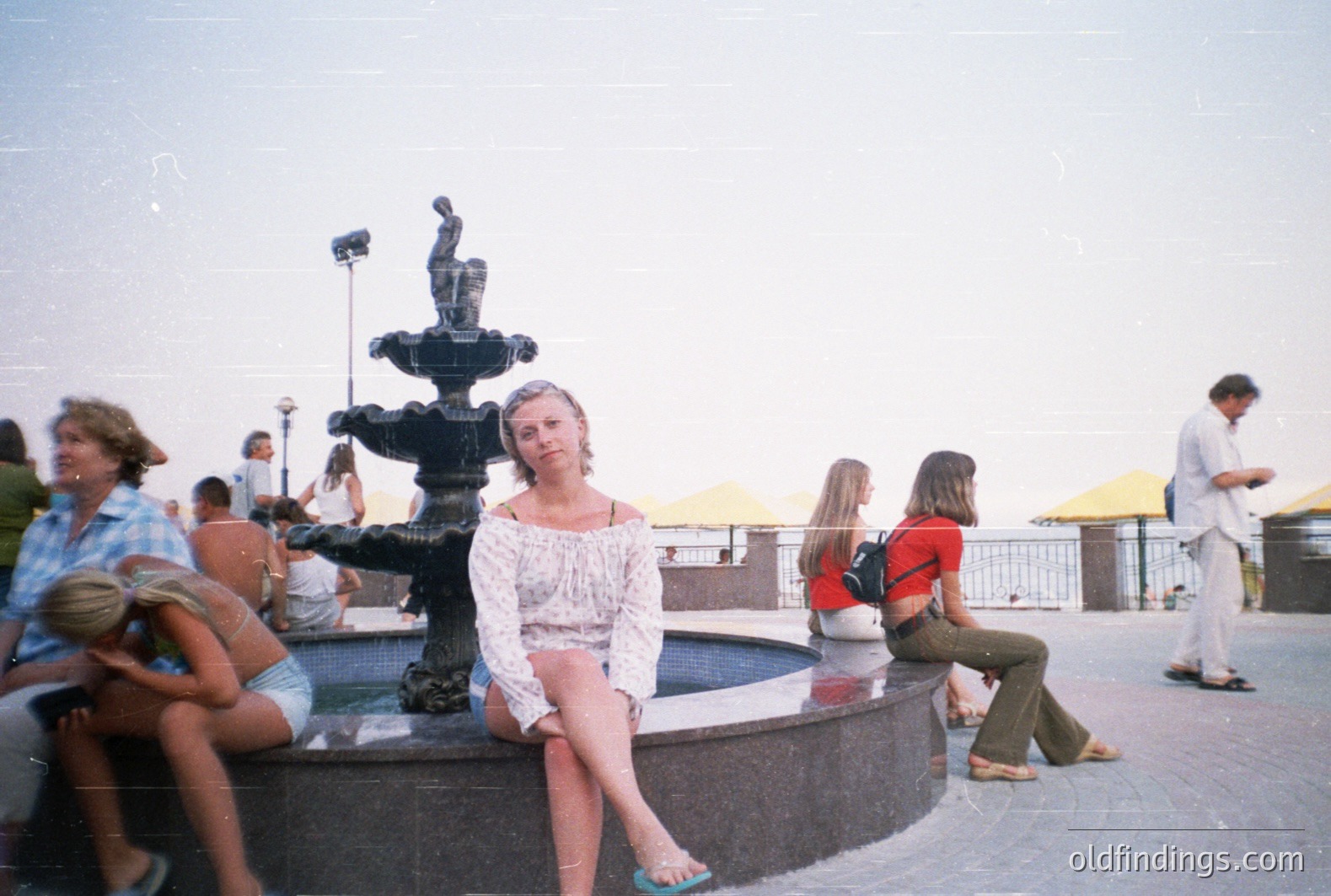 A young woman seated on a fountain's edge, posing for a candid snapshot. Figures populate a tiled plaza featuring a tiered fountain, overlooking a seaside view with distant yellow umbrellas. Likely taken in the 1970s or early 80s, perhaps in Bulgaria or a similar Eastern European resort. Interesting style reference.