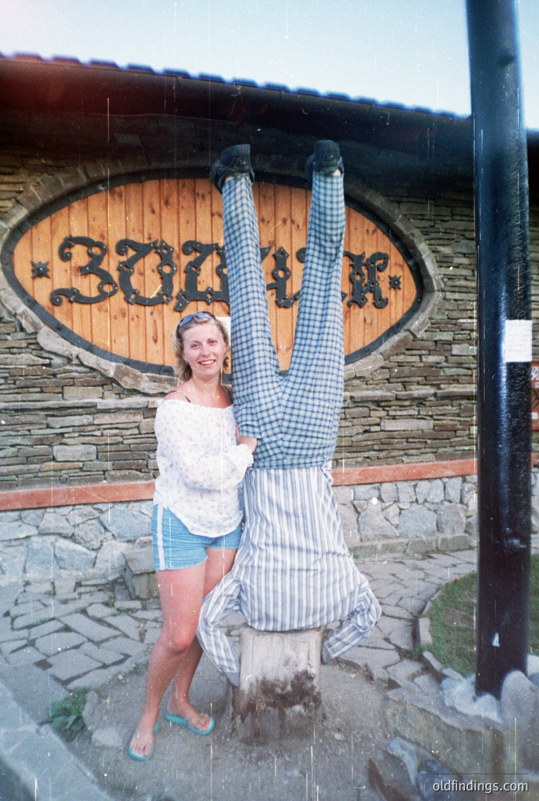 A woman in a white top and blue shorts poses beside a playful, oversized, cartoon-like figure with large eyes and a plaid shirt. The figure is suspended from a tree stump. Appears to be a tourist attraction or roadside novelty. Location likely coastal, judging by the terrain. Likely 1970s-80s era.