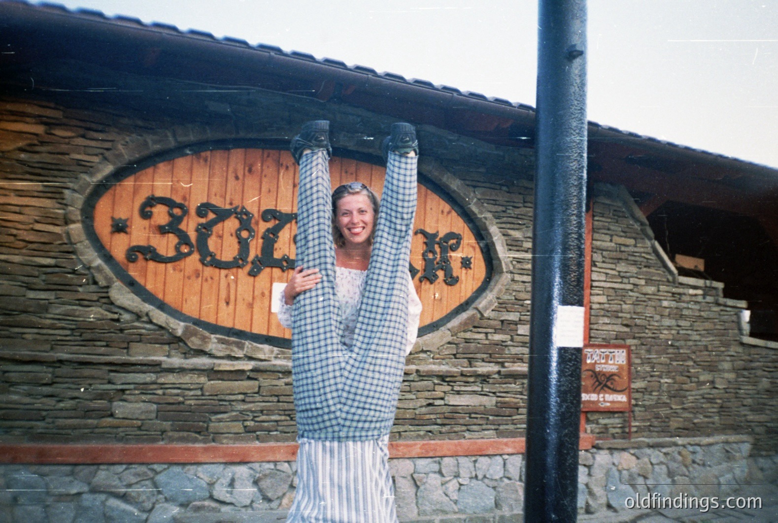 A man hangs upside down, wearing a blue and white striped shirt, in front of a stone building with an oval sign reading “302”. Likely a tourist photo, capturing a playful moment. Possible seaside resort architecture, 1970s style. Location uncertain but may be Eastern Europe.