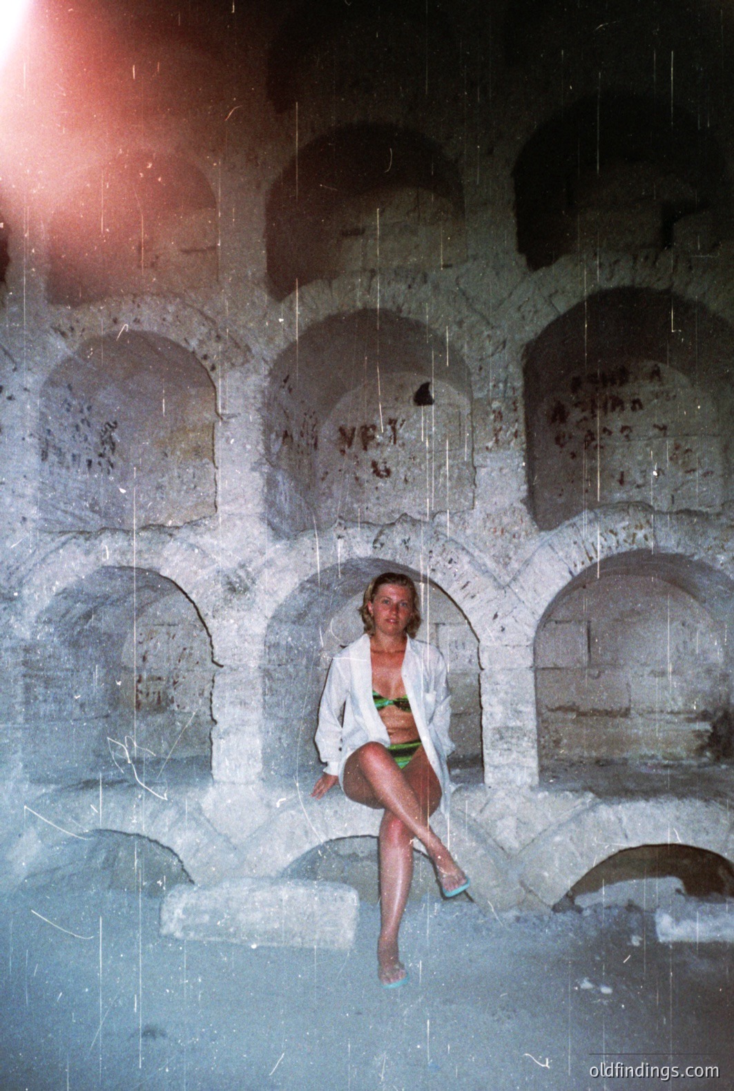 A woman in a swimsuit and white cover-up is seated within a stone archway structure, likely ruins or an old building. The architecture features a series of rounded arches with visible weathering and graffiti. Likely a tourist photo from the 1970s or 80s. Vintage film aesthetic.