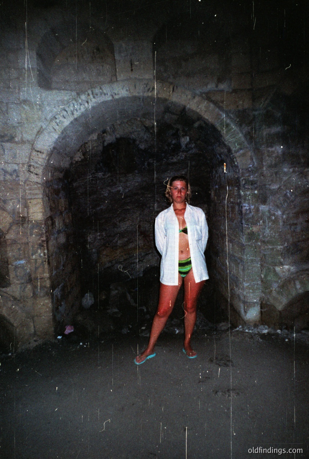 A woman stands beneath a stone archway, seemingly in the rain. She wears a bikini and an open white shirt, with flip-flops on her feet. The stone structure features rough-hewn blocks, creating a rustic, arched tunnel. The scene appears damp and likely coastal. Appears to be a candid, 1970s-era snapshot.