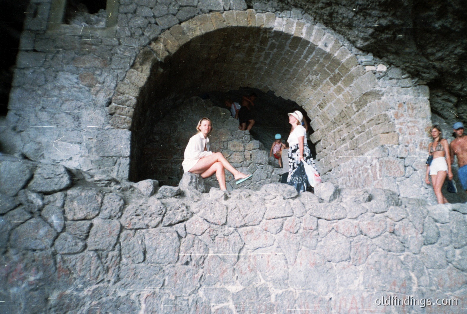Three young women are seen within a stone archway, seemingly part of a coastal structure. One woman sits, while the others stand, all appearing in swimwear. The archway is constructed of rough-hewn stone and brick. Likely a tourist location, 1970s aesthetic. Potential for travel/heritage stock use.