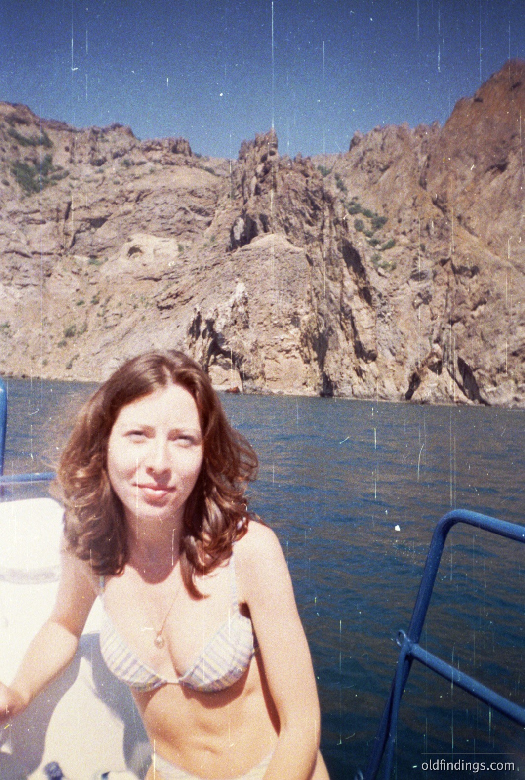A woman with shoulder-length dark hair sits on a boat, wearing a patterned bikini and a delicate necklace. Dramatic, rugged cliffs rise steeply from the tranquil water behind her. Likely a coastal scene – possibly Bulgaria's Black Sea coast. Mid-1970s color film aesthetic. A potential stock image representing leisure travel.
