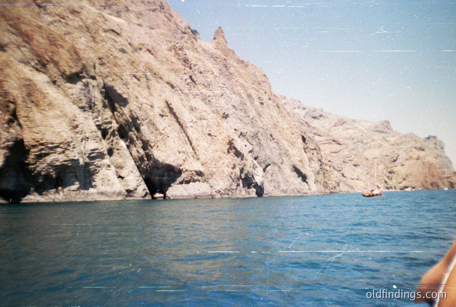 Dramatic coastal cliffs rise sharply from turquoise waters. A small boat is visible near the base of the rock face, emphasizing the scale. Likely Mediterranean coastline, possibly Bulgaria, given the rock formations. Appears to be a scan of a 1970s-era color print. Commercial value: scenic stock imagery.