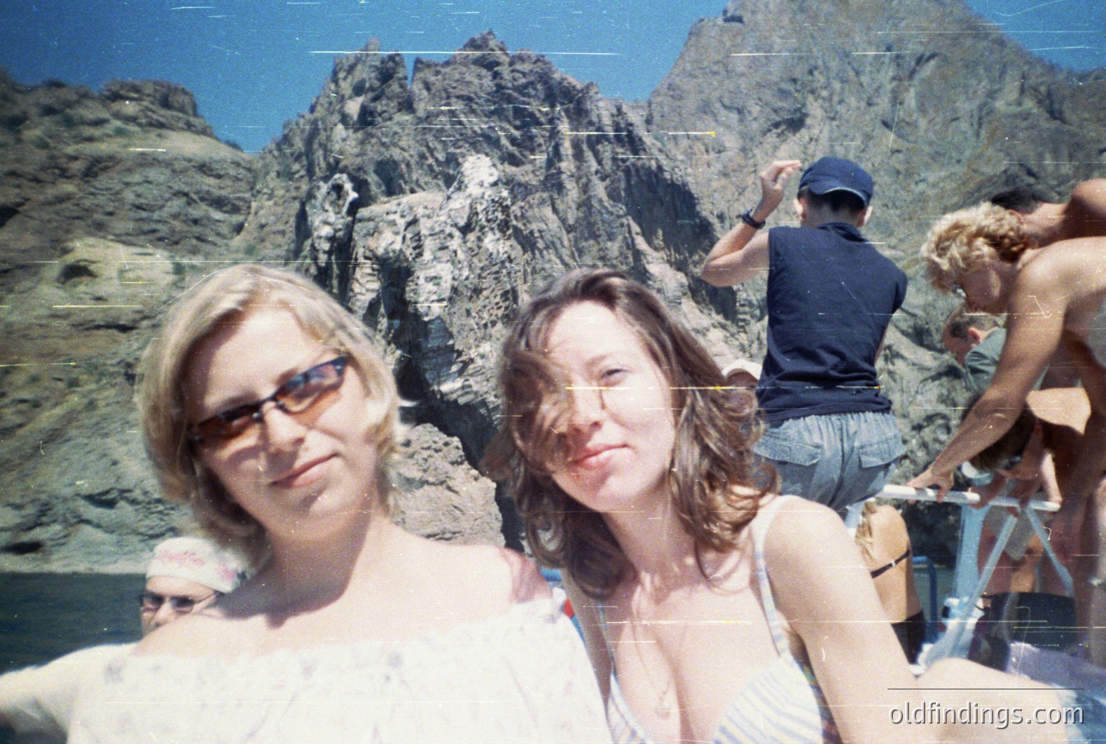 Two young women, one wearing sunglasses, pose outdoors with a rugged, rocky mountain backdrop. Several other figures are partially visible, suggesting a boat tour. The image's color palette and clothing styles indicate the 1990s. Likely a travel or vacation photograph.