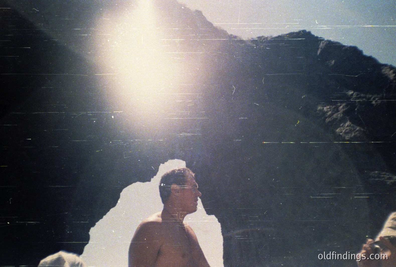A man in a short-sleeved shirt stands in profile, backlit by bright sunlight over a mountainous, rocky landscape. Likely a tourist snapshot from the 1970s or 80s, showcasing an alpine or coastal setting. Sun flares and film grain indicate analog photography. Potential commercial relevance for travel design or vintage stock.