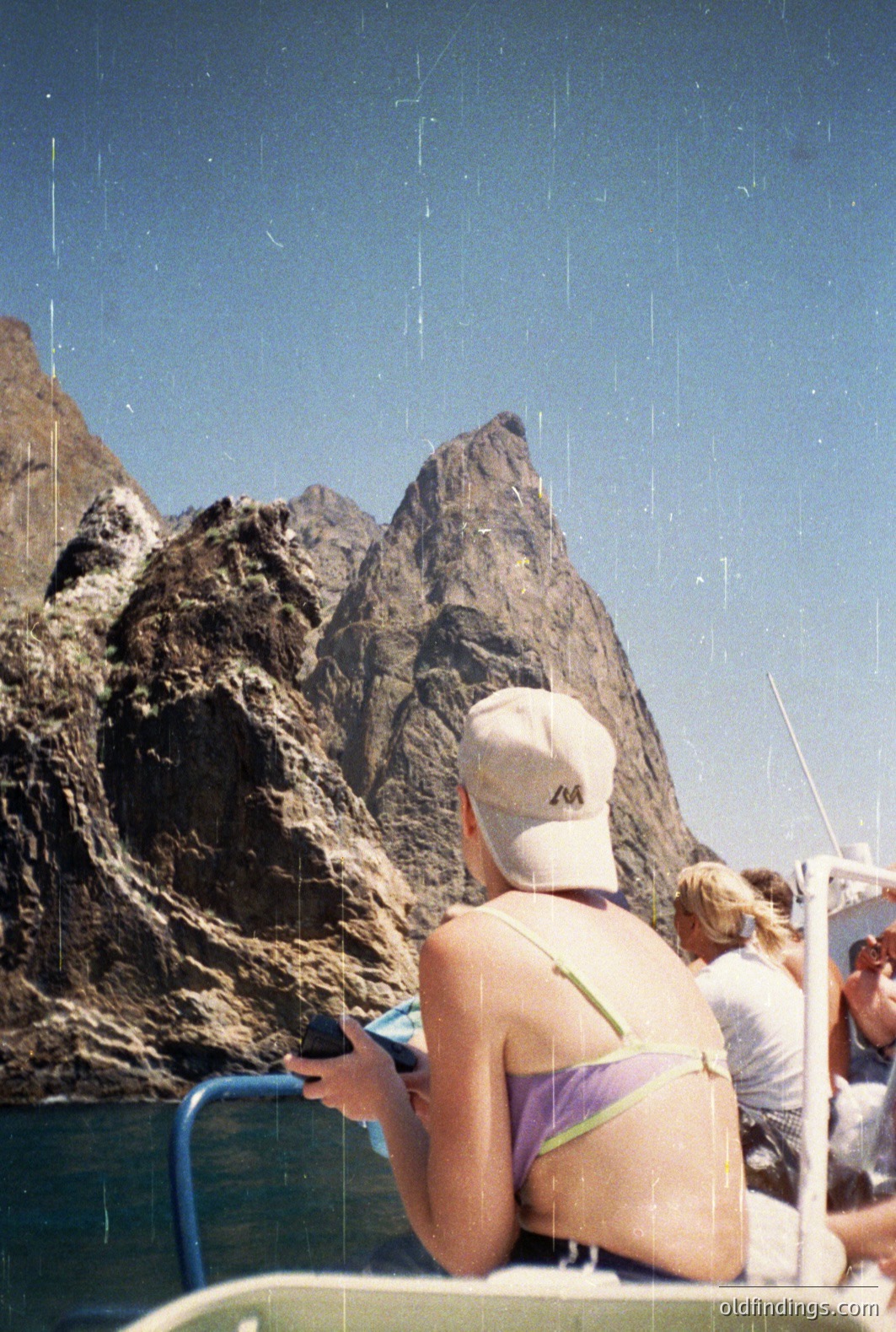A person in a purple bikini and cap is looking at a phone on a boat, with a dramatic, rocky mountain range visible beyond. The image exhibits a grainy texture, indicative of film photography. Likely coastal environment, possibly Mediterranean. Appears to be a candid moment.