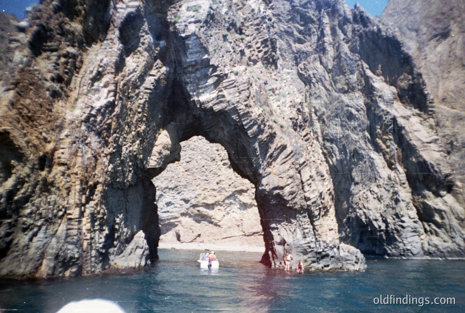 Dramatic coastal rock formation with a natural archway. Boats and figures visible near the shore within the arch. Heavily textured, layered rock faces suggest volcanic origin. Likely a seaside location, possibly Mediterranean. The image's color palette and composition evoke the 1970s-80s era.