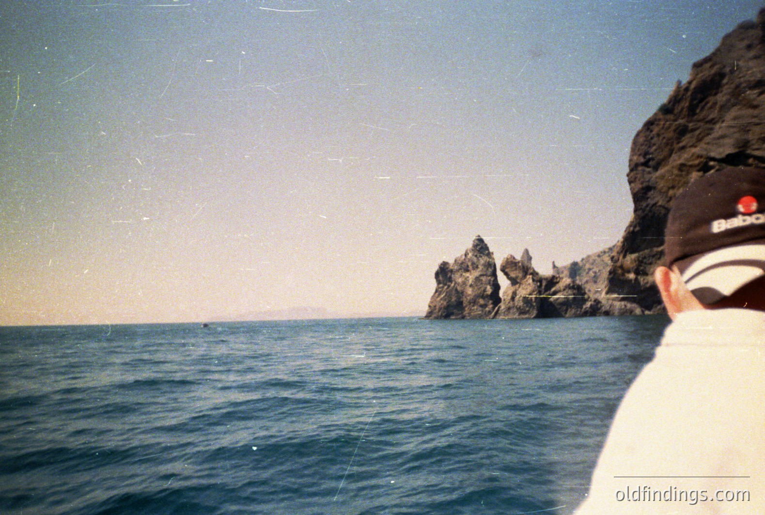 View from the sea towards rocky cliffs & a distant shoreline. A person wearing a Babolat visor and dark clothing occupies the foreground. The photo has a faded, vintage aesthetic. Likely captured in the 1980s or 1990s, possibly the Black Sea region.