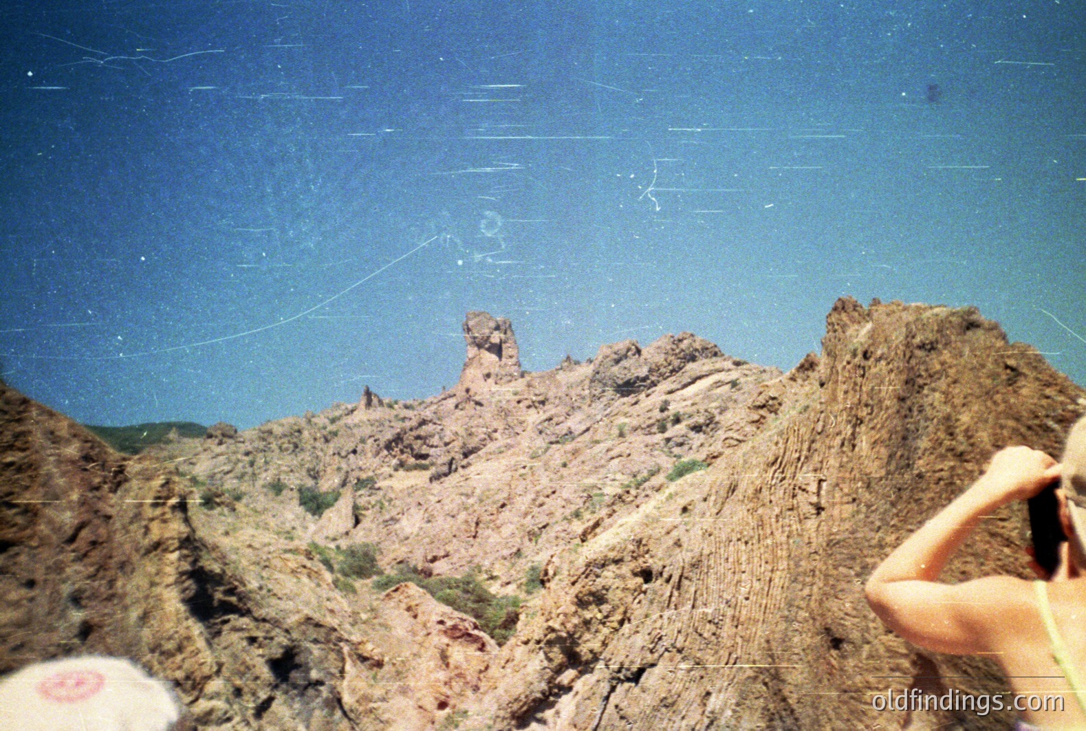 Striking, eroded sandstone rock formations dominate the scene against a bright blue sky. A person’s arm and hand, holding sunglasses, appear in the lower right corner. Visible texture suggests arid climate & geological processes. Likely Southwestern US location. Potential stock photo for travel or landscape design.