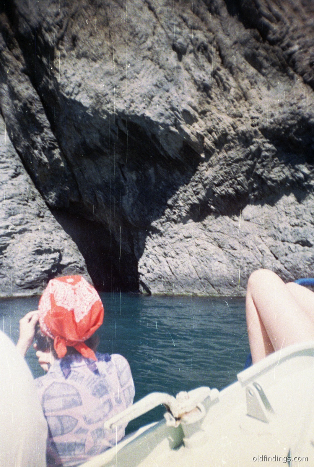 A person in a small boat navigates turquoise waters past a dramatic, rocky cliff face featuring a dark cave entrance. The individual wears a red bandana and patterned shirt. Likely coastal Mediterranean setting, potentially 1970s film aesthetic. A nostalgic, travel scene.